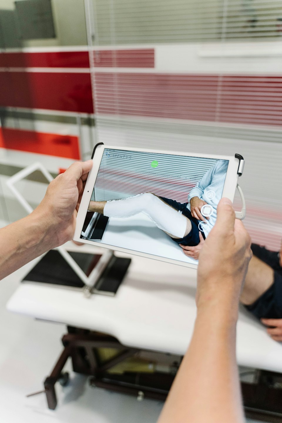 Hände halten ein Tablet, das ein Patientenbein auf einem Untersuchungstisch zeigt.